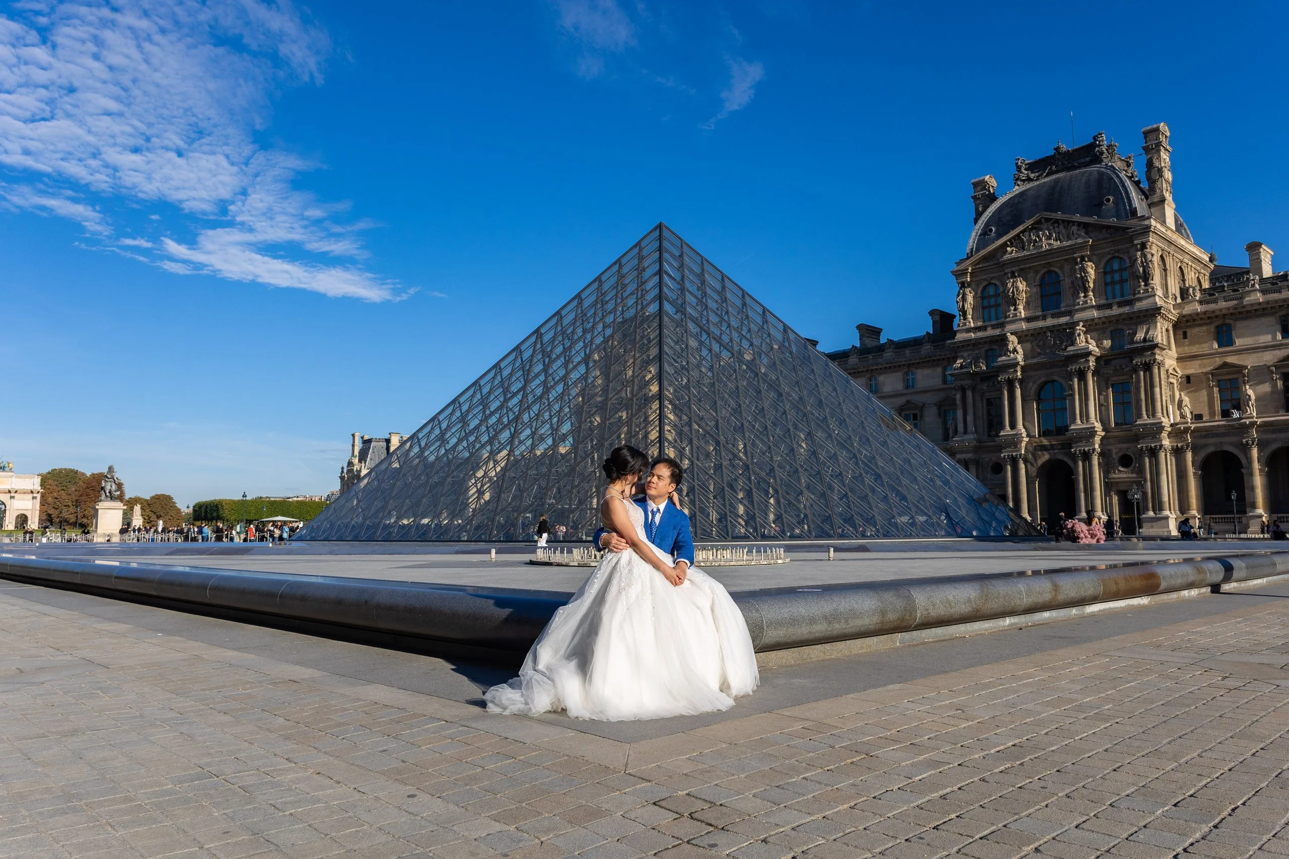 Mariés enlacés devant la pyramide du Louvre sous un ciel bleu, séance Day After à Paris