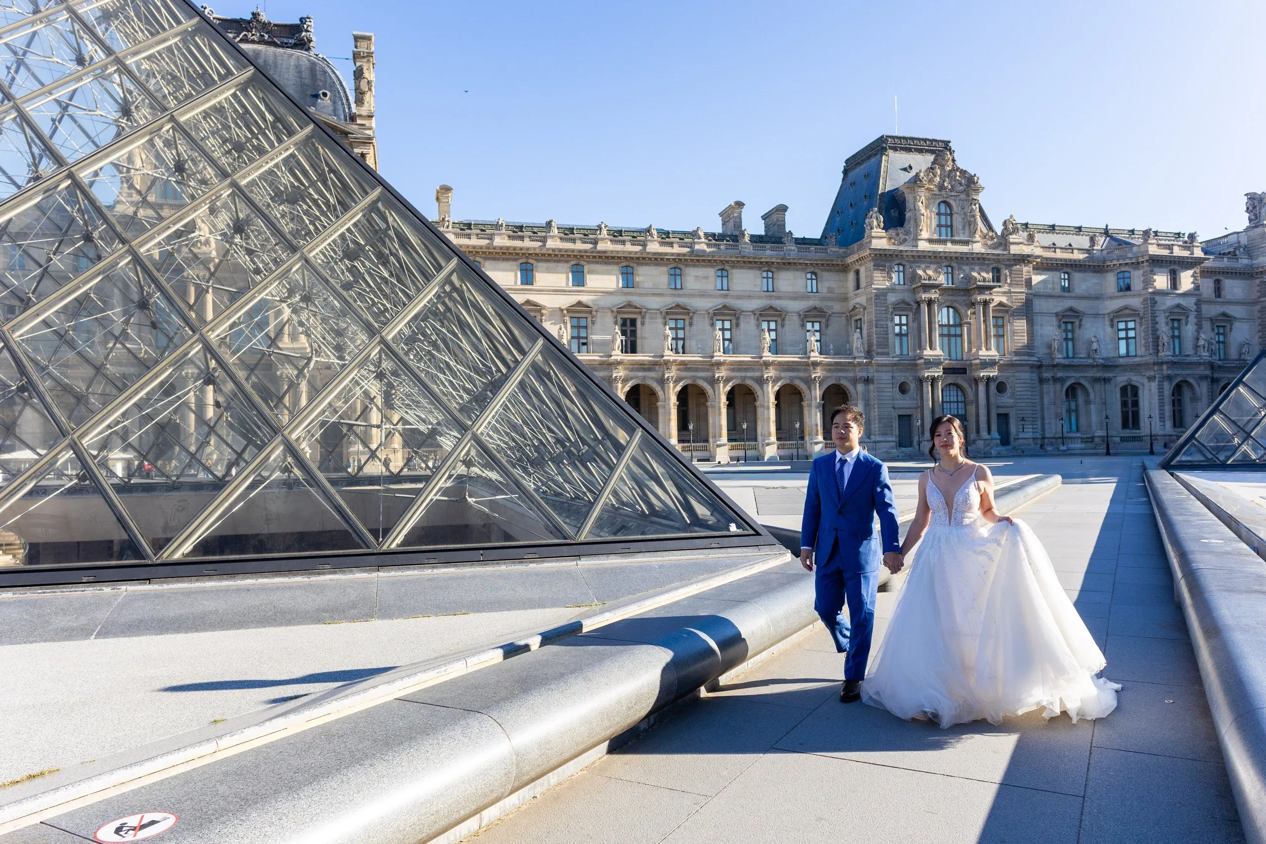 Couple de mariés marchant main dans la main devant la pyramide du Louvre